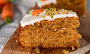 Ein saftiges Stück Karottenkuchen aus der Kastenform, dekoriert mit weißem Zuckerguss, gehackten Pistazien und einer kleinen Marzipankarotte.