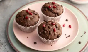 Drei saftige, vegane Muffins mit Schokostückchen und Granatapfelkernen auf einem rosa Teller.
