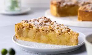 Ein saftiges Stück veganer Apfelkuchen mit Streuseln und Puderzucker bestäubt. Der Kuchen liegt auf einem weißen Teller, im Hintergrund ist ein ganzer Kuchen zu sehen.