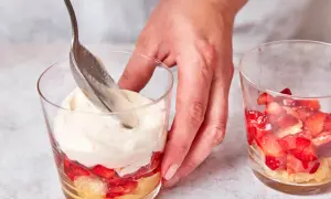 Eine Hand schichtet mit einem Löffel cremiges Topping auf ein Erdbeer-Tiramisu im Glas.