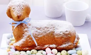 Ein süßes Osterlamm-Rezept: Ein goldbrauner Kuchen in Lammform, mit Puderzucker bestäubt und einem blauen Band verziert, umgeben von bunten Zuckereiern.
