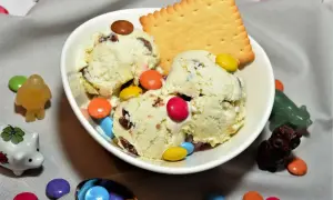 Schüssel mit cremigem Eis, garniert mit bunten Smarties und einem Keks. Kleine Figuren und weitere Smarties schmücken den Tisch.