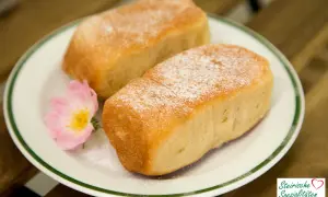 Zwei goldbraune Buchteln, mit Puderzucker bestäubt, auf einem Teller mit einer rosa Blüte. Ein köstliches Buchteln Rezept für Genießer.