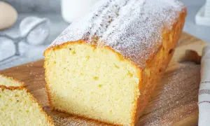 Ein einfacher Kuchen, bestäubt mit Puderzucker, liegt auf einem Holzbrett. Daneben liegen zwei Scheiben des Kuchens.