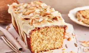 Ein saftiger Kokos Kuchen mit weißer Glasur und gerösteten Kokoschips. Daneben liegt eine Kokosnuss.