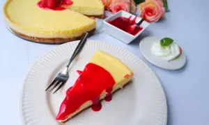 Ein Stück New York Cheesecake mit roter Fruchtsauce und Himbeeren, daneben eine kleine Schüssel Sauce und ein Klecks Sahne.