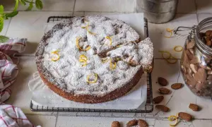 Ein mallorquinischer Mandelkuchen, bestäubt mit Puderzucker und garniert mit Zitronenzesten, neben einem Glas Mandeln.