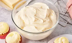 Schüssel mit cremiger amerikanischer Buttercreme, daneben ein Block Butter, Cupcakes und ein Handmixer.