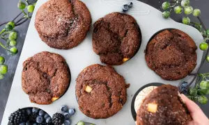 Fünf schoko muffins mit weißen Schokostückchen und Puderzucker in einer Muffinform, daneben Beeren und grüne Beeren.