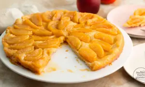 Eine goldbraune Tarte Tatin mit karamellisierten Apfelspalten auf einem weißen Teller. Ein Stück fehlt, was Appetit macht.