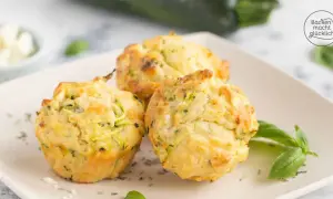 Drei goldbraune Zucchini-Muffins mit Kräutern und Käse auf einem Teller, garniert mit Basilikum.
