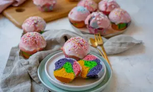 Bunte regenbogen muffins mit rosa Zuckerguss und Streuseln, ein Stück auf einem Teller zeigt das farbenfrohe Innere.