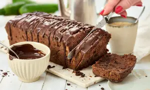 Ein saftiger Zucchini Schoko Kuchen wird angeschnitten. Daneben eine Schale mit Schokoladenglasur und eine Tasse Kaffee.