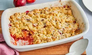 Ein frisch gebackener pflaumen crumble in einer weißen Auflaufform, bereit zum Servieren. Daneben liegen zwei reife Pflaumen.