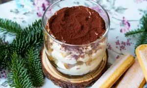 Ein köstliches Tiramisu im Glas, bestäubt mit Kakao, neben Löffelbiskuits und Tannenzweigen.