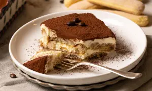 Ein Stück Tiramisu, das nach einem einfachen tiramisu rezept gemacht wurde, liegt auf einem Teller. Kakaopulver bestäubt die cremige Schicht und die Löffelbiskuits.