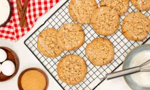 Frisch gebackene Zimt-Streusel-Kekse auf einem Kuchengitter. Zutaten wie Eier, Zucker und Mehl sind daneben zu sehen.