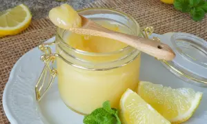 Ein Glas mit cremigem Lemon Curd, garniert mit Zitronenspalten und Minze. Ein Holzlöffel mit einer Portion Lemon Curd ruht auf dem Glas.