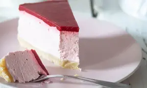Ein Stück cremiger cheesecake ohne backen mit roter Fruchtschicht auf rosa Teller, daneben ein Gabel mit einem Stück Kuchen.