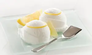 Zitroneneis selber machen ist einfach! Zwei Kugeln erfrischendes Zitroneneis mit Zitronenspalten und einem Eisportionierer auf einem Glas-Tablett.