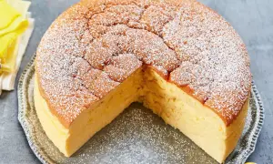 Ein fluffiger, goldbrauner low carb kuchen, bestäubt mit Puderzucker, auf einer silbernen Platte. Ein kleines gelbes Vogel-Dekoration sitzt daneben.