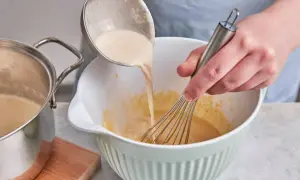 Eine cremige Masse wird aus einem Topf in eine Schüssel gegossen, während mit einem Schneebesen gerührt wird. Perfekt für selbstgemachtes Zimteis aus der Eismaschine.