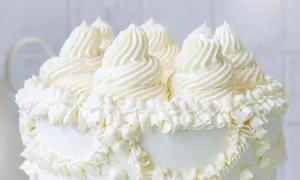 Eine üppige Buttercreme Torte mit kunstvollen Tupfen und Rüschen aus weißer Buttercreme, die auf einem weißen Hintergrund steht.