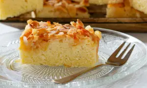 Ein saftiges Stück mandelkuchen vom blech, bestreut mit gerösteten Mandeln und Zucker, liegt auf einem Glas-Teller.