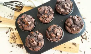 Sechs saftige, vegane Schokomuffins mit Schokostückchen in einer Muffinform. Ein Schneebesen mit Schokospuren liegt daneben.