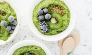 Grüne nice cream mit Kiwi, Beeren und Brombeere in weißen Schalen, daneben Holzlöffel.