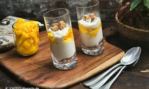 Zwei Gläser mit einem cremigen Mango-Dessert, geschichtet mit zerbröselten Keksen und garniert mit Mango-Stücken. Daneben ein Glas voll frischer Mango-Würfel.