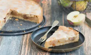 Ein Stück saftiger Apfelkuchen mit süßer Glasur und Mandelsplittern, daneben ein halber Apfel und ein Korb voller Äpfel.
