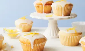 Saftige vegane Zitronenmuffins mit Zuckerguss und Zitronenschale verziert, auf einem weißen Kuchenständer und Tisch.