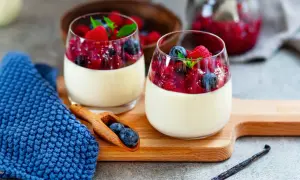 Zwei Gläser cremiger Panna cotta mit Beerenkompott und frischen Beeren, garniert mit Minze. Ein Löffel mit Blaubeeren liegt daneben.