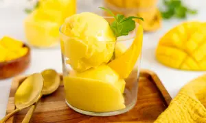 Erfrischendes Mango Eis in einem Glas, garniert mit Minze und Mangostücken. Daneben liegen goldene Löffel auf einem Holztablett.