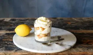 Ein Glas mit geschichtetem Zitronen Tiramisu, garniert mit Zitronenschale. Daneben liegt eine Zitrone.
