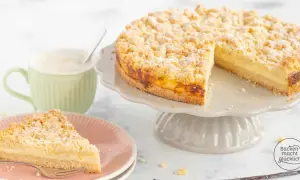 Ein saftiger Streuselkuchen mit Puddingfüllung, bestäubt mit Puderzucker. Daneben eine Tasse Kaffee und ein Stück Kuchen auf einem Teller.