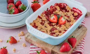 Ein frisch gebackener Erdbeer Crumble in einer hellblauen Auflaufform, garniert mit Erdbeerhälften. Frische Erdbeeren liegen daneben.