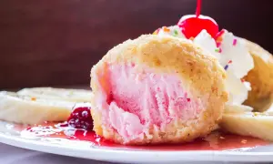 Ein knusprig gebratenes Eis mit rosa Füllung, garniert mit Sahne, Kirsche und bunten Streuseln. Dazu Bananenscheiben und rote Sauce.