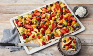 Bunter Blechkuchen mit Obst, belegt mit Erdbeeren, Kiwi, Trauben, Pfirsichen und Beeren. Ein Stück ist bereits auf einem Teller angerichtet.