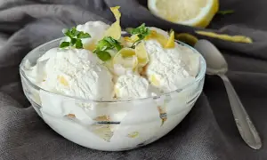 Ein Glas cremiges zitronen eis, garniert mit Minze und Zitronenschale, daneben eine halbe Zitrone und ein Löffel.