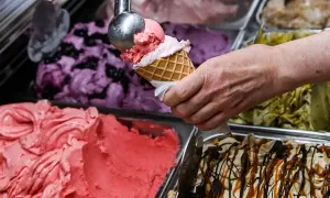Eine Hand füllt eine Waffel mit rosa Eis. Daneben stehen Behälter mit verschiedenen Eissorten, darunter Blaubeer- und Pistazieneis. Das beste Eis der Welt!