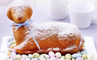 Ein süßes Osterlamm-Rezept: Ein goldbrauner Kuchen in Lammform, mit Puderzucker bestäubt und einem blauen Band verziert, umgeben von bunten Zuckereiern.