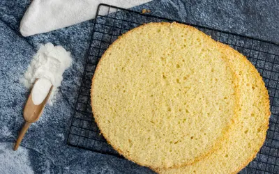 Zwei runde, goldgelbe Scheiben eines luftigen, **glutenfreien Biskuitbodens** liegen auf einem schwarzen Gitter. Daneben ein Holzlöffel mit weißem Pulver.