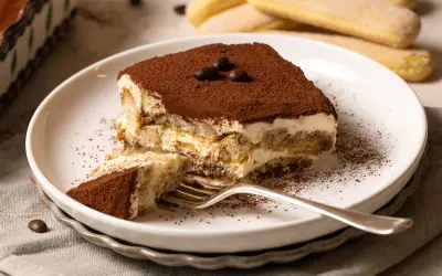 Ein Stück Tiramisu, das nach einem einfachen tiramisu rezept gemacht wurde, liegt auf einem Teller. Kakaopulver bestäubt die cremige Schicht und die Löffelbiskuits.