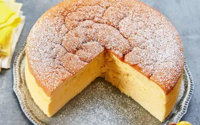 Ein fluffiger, goldbrauner low carb kuchen, bestäubt mit Puderzucker, auf einer silbernen Platte. Ein kleines gelbes Vogel-Dekoration sitzt daneben.