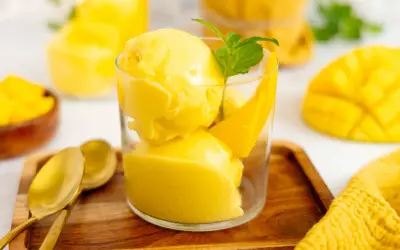 Erfrischendes Mango Eis in einem Glas, garniert mit Minze und Mangostücken. Daneben liegen goldene Löffel auf einem Holztablett.