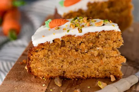 Ein saftiges Stück Karottenkuchen aus der Kastenform, dekoriert mit weißem Zuckerguss, gehackten Pistazien und einer kleinen Marzipankarotte.