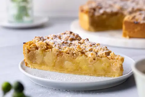 Ein saftiges Stück veganer Apfelkuchen mit Streuseln und Puderzucker bestäubt. Der Kuchen liegt auf einem weißen Teller, im Hintergrund ist ein ganzer Kuchen zu sehen.