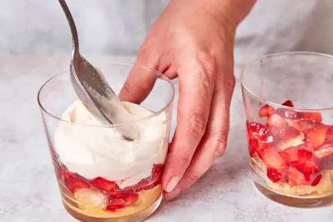 Eine Hand schichtet mit einem Löffel cremiges Topping auf ein Erdbeer-Tiramisu im Glas.